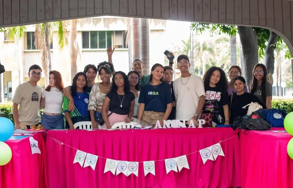 Un grupo grande de estudiantes frente a un stand con mantel fucsia.